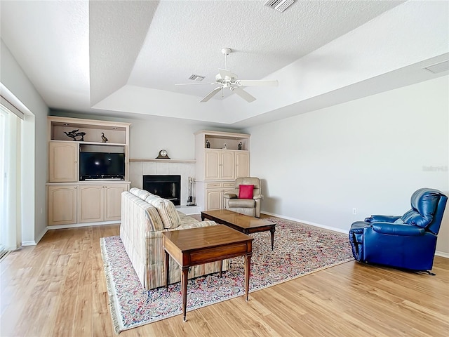 living room featuring light wood-type flooring, a tiled fireplace, ceiling fan, a raised ceiling, and a textured ceiling