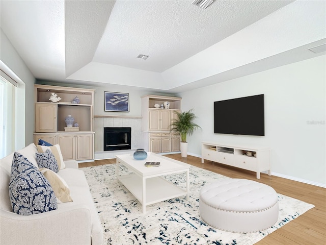 living room with a tiled fireplace, light hardwood / wood-style floors, a raised ceiling, and a textured ceiling