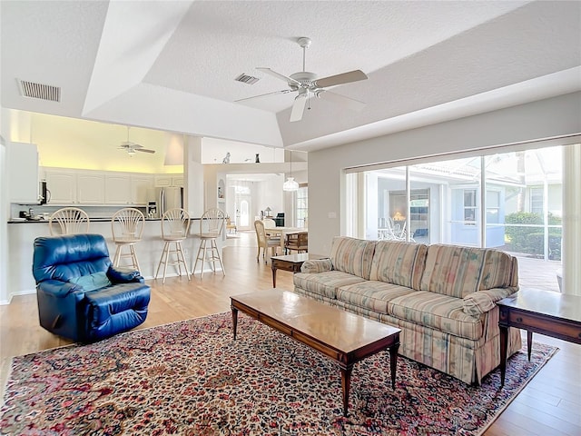 living room with lofted ceiling, a textured ceiling, light wood-type flooring, a raised ceiling, and ceiling fan