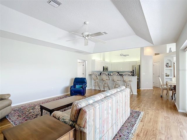 living room featuring built in shelves, a textured ceiling, light wood-type flooring, a raised ceiling, and ceiling fan