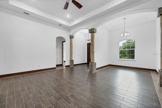 empty room with decorative columns, ceiling fan with notable chandelier, dark hardwood / wood-style floors, and a tray ceiling