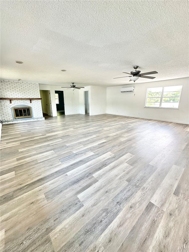 unfurnished living room featuring a textured ceiling, a wall mounted air conditioner, a brick fireplace, ceiling fan, and light wood-type flooring