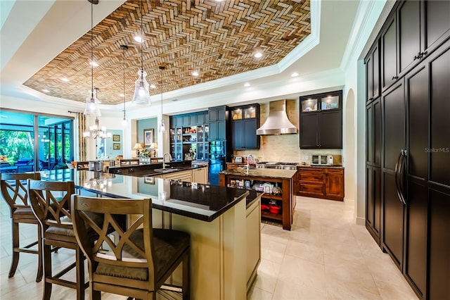 kitchen featuring wall chimney exhaust hood, a kitchen island with sink, a kitchen breakfast bar, and a tray ceiling