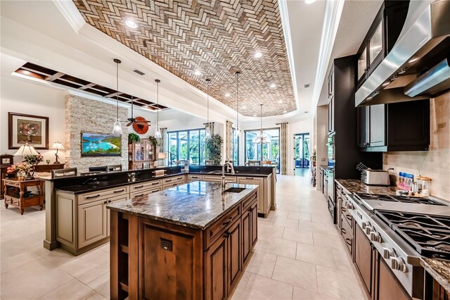 kitchen featuring wall chimney range hood, dark stone countertops, a raised ceiling, and a large island with sink