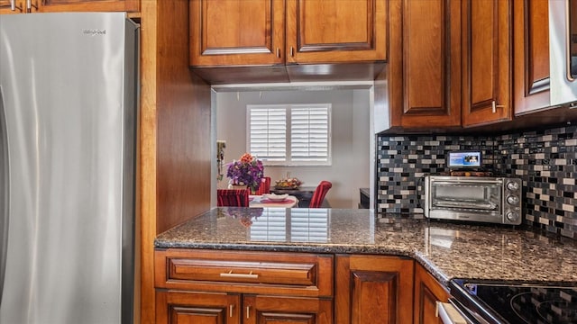 kitchen featuring dark stone counters, tasteful backsplash, brown cabinetry, a toaster, and freestanding refrigerator