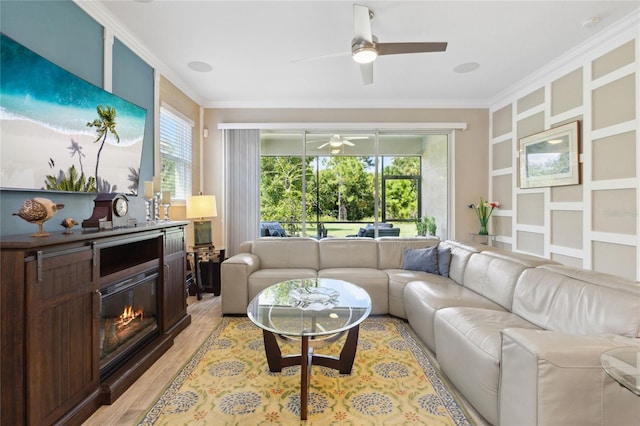 living room with ceiling fan, ornamental molding, a glass covered fireplace, and light wood-style floors