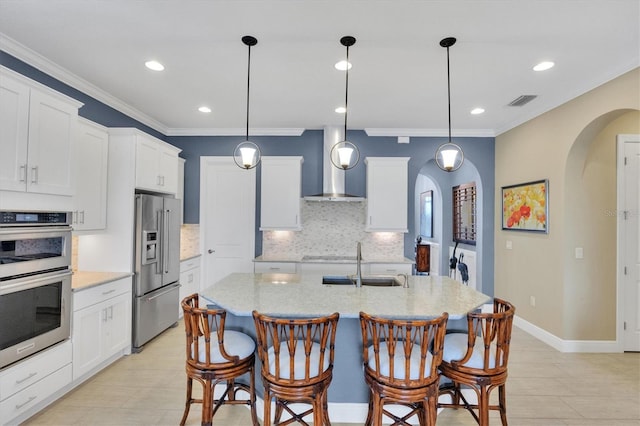 kitchen with wall chimney exhaust hood, an island with sink, appliances with stainless steel finishes, and hanging light fixtures