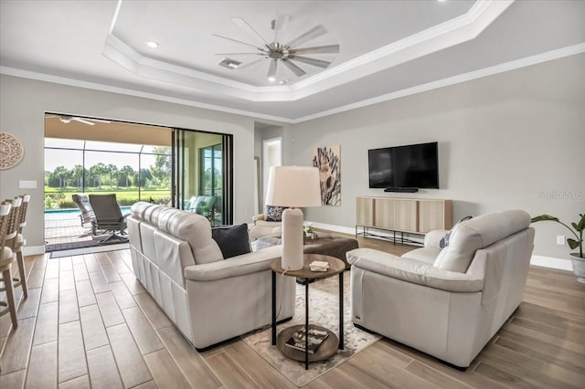 living room featuring ceiling fan, ornamental molding, a tray ceiling, and light wood-type flooring