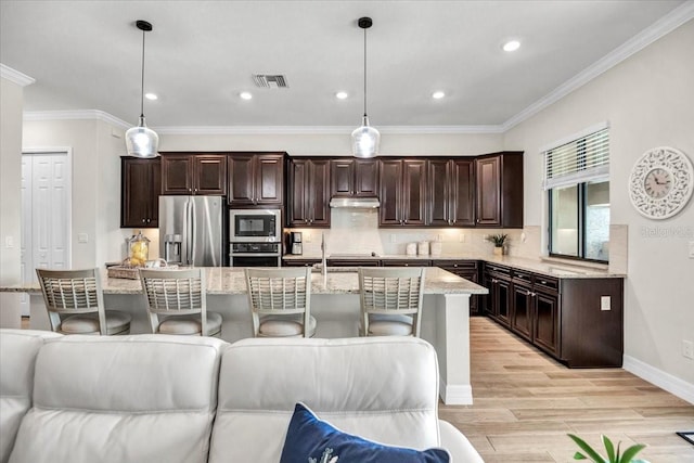 kitchen featuring tasteful backsplash, light wood-type flooring, an island with sink, hanging light fixtures, and stainless steel appliances