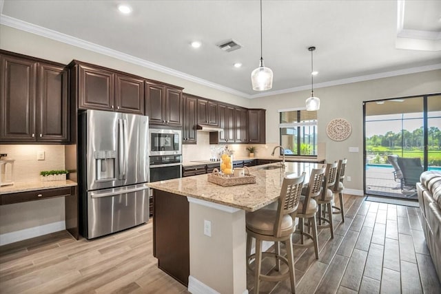 kitchen with light hardwood / wood-style floors, stainless steel appliances, dark brown cabinetry, pendant lighting, and a center island with sink