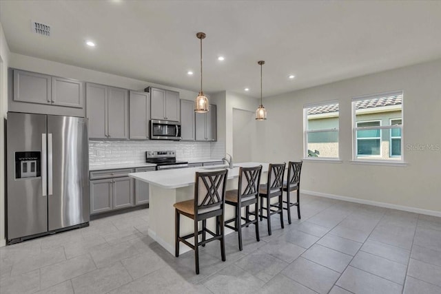 kitchen featuring gray cabinetry, a kitchen island with sink, appliances with stainless steel finishes, decorative light fixtures, and a breakfast bar area