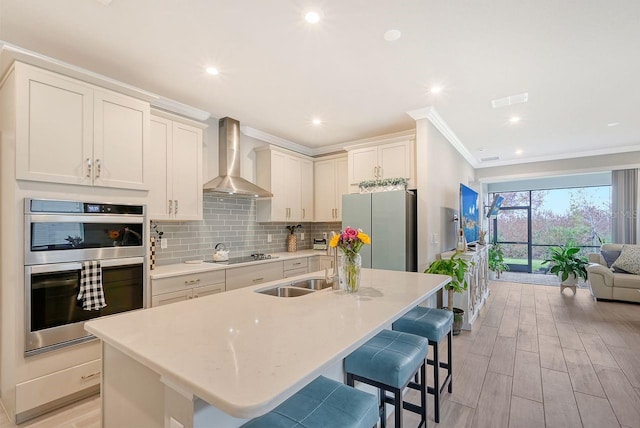 kitchen with wall chimney range hood, an island with sink, a breakfast bar, stainless steel appliances, and light hardwood / wood-style flooring