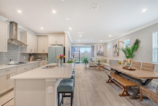 kitchen featuring wall chimney range hood, stainless steel fridge, an island with sink, light hardwood / wood-style flooring, and sink