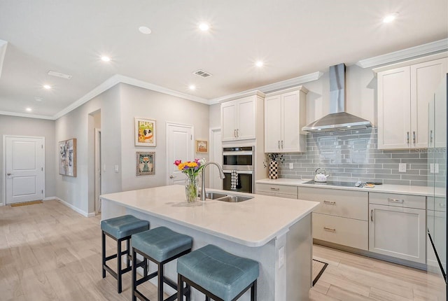 kitchen with sink, light wood-type flooring, an island with sink, double oven, and wall chimney exhaust hood