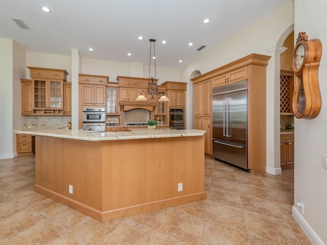 kitchen featuring light stone counters, backsplash, appliances with stainless steel finishes, a large island with sink, and decorative light fixtures