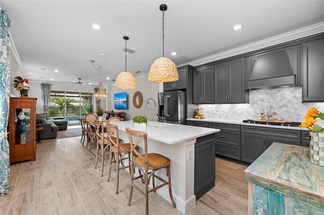 kitchen featuring custom exhaust hood, an island with sink, appliances with stainless steel finishes, ornamental molding, and pendant lighting