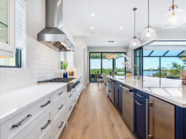 1109 Bayshore Rd, Nokomis, FL 34275 - kitchen featuring wall chimney exhaust hood, blue cabinets, appliances with stainless steel finishes, light wood-style flooring, and tasteful backsplash