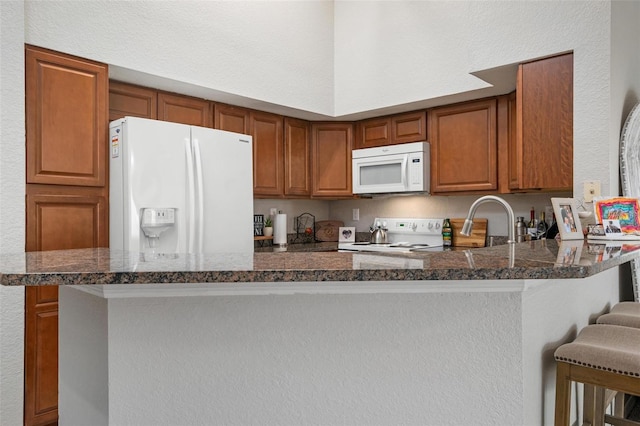 kitchen featuring white appliances, sink, kitchen peninsula, dark stone counters, and a breakfast bar