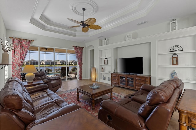 tiled living area featuring built in features, a raised ceiling, a ceiling fan, arched walkways, and crown molding