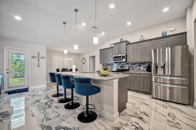 kitchen featuring stainless steel appliances, hanging light fixtures, a breakfast bar area, a kitchen island with sink, and light stone countertops