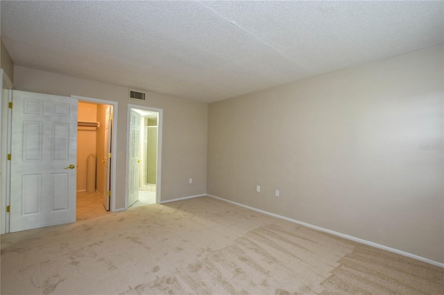 unfurnished bedroom with ensuite bathroom, a walk in closet, a textured ceiling, light colored carpet, and a closet