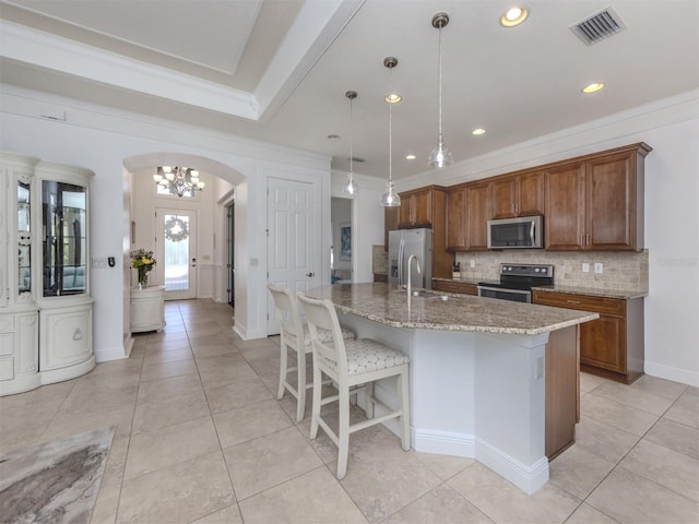 kitchen featuring appliances with stainless steel finishes, backsplash, light stone counters, decorative light fixtures, and an island with sink