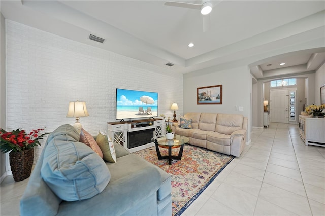 tiled living room featuring ceiling fan, a raised ceiling, and brick wall