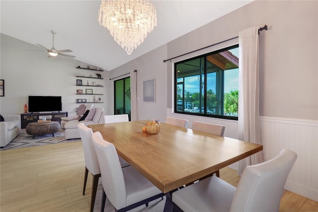 dining room with light wood finished floors, ceiling fan with notable chandelier, wainscoting, and vaulted ceiling