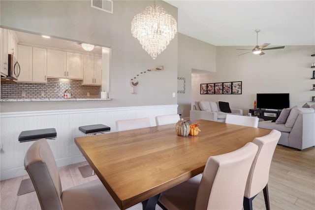 dining space featuring a wainscoted wall, visible vents, light wood finished floors, a towering ceiling, and ceiling fan with notable chandelier