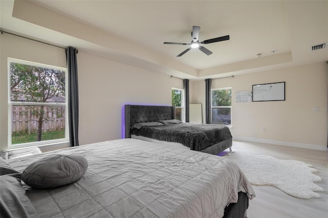 bedroom with ceiling fan, light wood-type flooring, a tray ceiling, and multiple windows