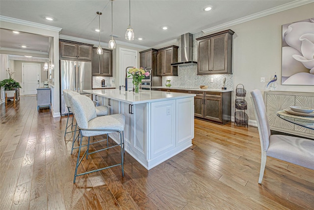 kitchen featuring an island with sink, a breakfast bar area, hanging light fixtures, high end fridge, and wall chimney range hood