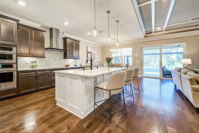 kitchen featuring a breakfast bar area, stainless steel double oven, a kitchen island with sink, dark brown cabinetry, and wall chimney range hood