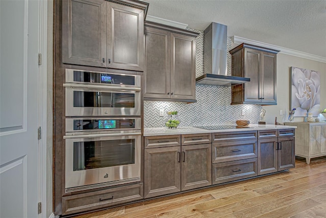 kitchen with wall chimney exhaust hood, dark brown cabinetry, tasteful backsplash, light wood-type flooring, and stainless steel double oven