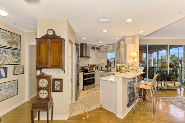 kitchen featuring wall chimney exhaust hood, a kitchen breakfast bar, light hardwood / wood-style flooring, kitchen peninsula, and stainless steel range with electric stovetop