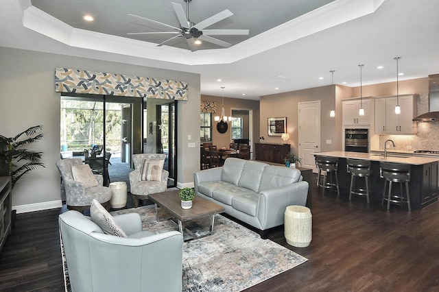 living room featuring ceiling fan with notable chandelier, a raised ceiling, crown molding, sink, and dark hardwood / wood-style floors