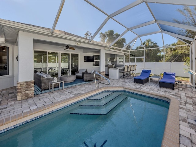 view of pool featuring outdoor lounge area, glass enclosure, ceiling fan, a patio area, and exterior kitchen