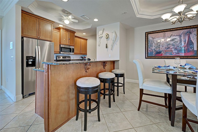 kitchen featuring stainless steel appliances, a raised ceiling, a kitchen breakfast bar, dark stone counters, and ceiling fan with notable chandelier