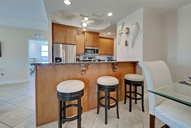 kitchen with a kitchen breakfast bar, ornamental molding, dark stone counters, stainless steel appliances, and a tray ceiling