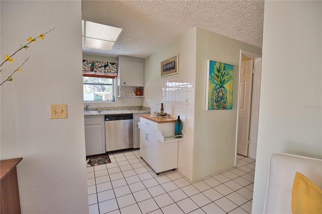 kitchen with stainless steel dishwasher, a textured ceiling, light tile patterned floors, tile walls, and white cabinetry