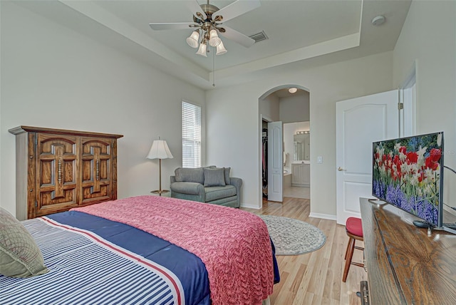 bedroom with light wood-type flooring, connected bathroom, ceiling fan, and a raised ceiling