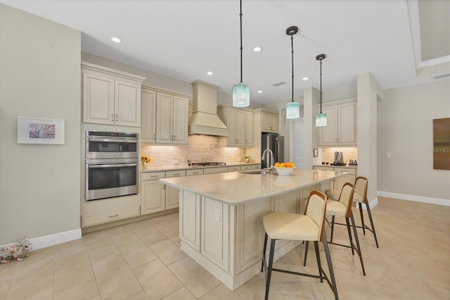 kitchen featuring appliances with stainless steel finishes, premium range hood, a kitchen island with sink, cream cabinetry, and hanging light fixtures