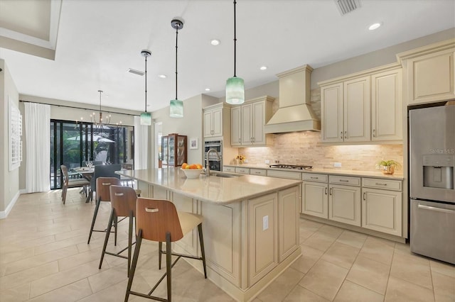 kitchen with appliances with stainless steel finishes, custom exhaust hood, cream cabinets, decorative light fixtures, and an island with sink