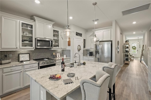 kitchen with hanging light fixtures, appliances with stainless steel finishes, glass insert cabinets, a kitchen island with sink, and a sink