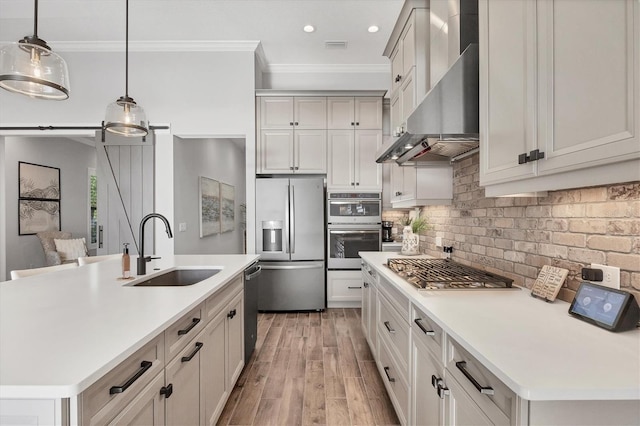 kitchen featuring appliances with stainless steel finishes, wall chimney exhaust hood, sink, decorative light fixtures, and an island with sink