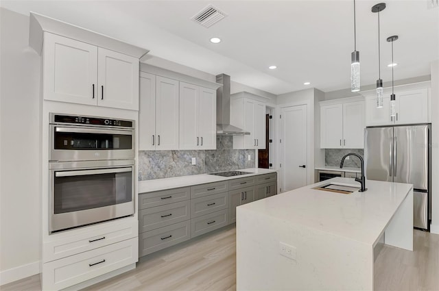 kitchen featuring appliances with stainless steel finishes, wall chimney exhaust hood, sink, a center island with sink, and decorative light fixtures