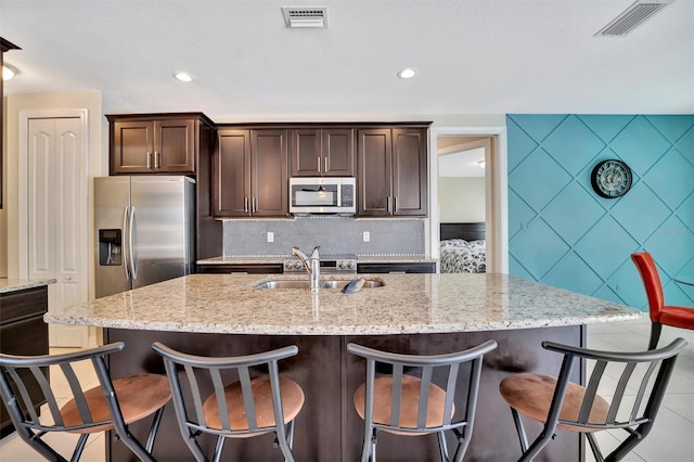 kitchen with light stone counters, dark brown cabinets, stainless steel appliances, a kitchen island with sink, and sink