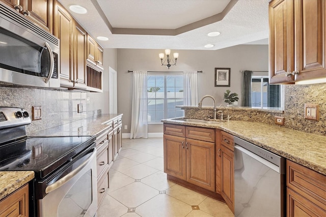 kitchen featuring a raised ceiling, appliances with stainless steel finishes, light stone countertops, a notable chandelier, and sink