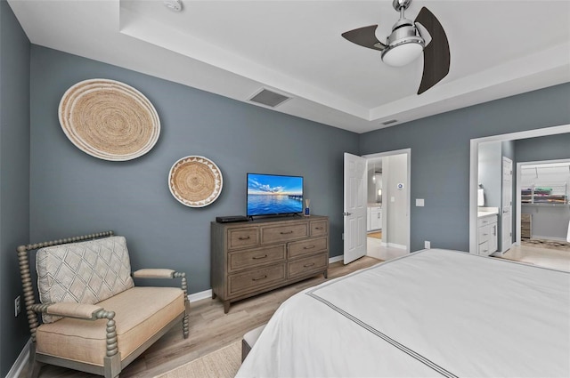 bedroom with light wood-type flooring, ensuite bathroom, ceiling fan, and a tray ceiling