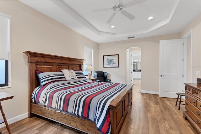 bedroom featuring ceiling fan, a raised ceiling, light wood-type flooring, and ensuite bath