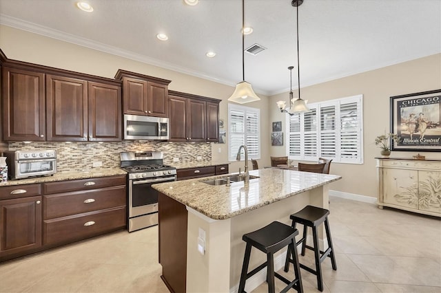 kitchen with appliances with stainless steel finishes, light stone counters, a kitchen island with sink, sink, and hanging light fixtures